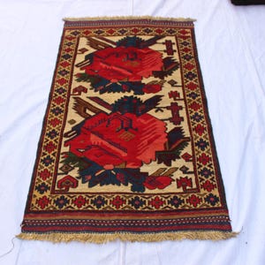 May include: Rectangular rug with a beige background and a large red floral design. The rug has a dark brown border with intricate patterns and a fringed edge. The floral design includes blue and green accents.