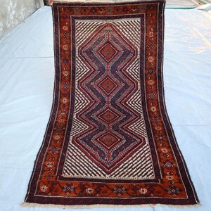 Puede incluir: Una alfombra rectangular larga con un patrón geométrico en tonos rojos, azules y blancos. La alfombra tiene un borde marrón con intrincados diseños florales.