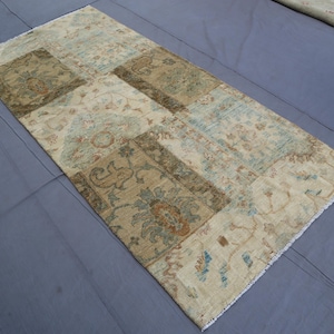 Puede incluir: Una alfombra rectangular con un diseño de patchwork compuesto por varias secciones con intrincados patrones florales y botánicos en tonos apagados de beige, marrón claro, verde oliva y azul claro, terminada con flecos blancos cortos en los extremos.