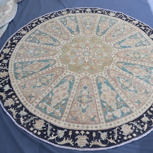 Könnte beinhalten: Ein runder Teppich mit einem aufwendigen Blumenmuster. Der Teppich hat ein zentrales Blumenmedaillon in Beige, Türkis und Braun, umgeben von einem dunkelblauen Rand mit floralen Motiven. Der Teppich hat einen Durchmesser von etwa 183 cm.