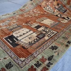 May include: Hand-knotted rug with a central design featuring stylized vases and floral motifs in shades of rust, beige, and black. The rug has a sage green border with repeating leaf patterns. The rug is rectangular.