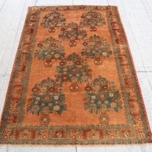 May include: An orange-toned rug with a floral design. The rug features multiple bouquets of flowers in vases, with a border of intricate patterns. The rug's color palette includes shades of orange, green, and brown.