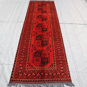 May include: A long rectangular runner rug featuring a vibrant red background with repeating black geometric motifs and stylized tree patterns. The border displays intricate black and red zigzag and floral designs, finished with grey fringe at both ends.