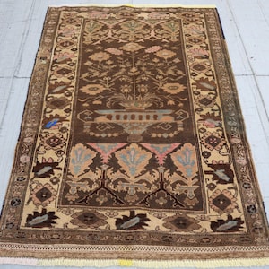 May include: A rectangular, hand-knotted rug with a central brown field featuring floral and geometric patterns in beige, pink, and blue. The rug has a beige border with intricate designs and a fringed edge. The rug measures approximately 91 x 152 cm.
