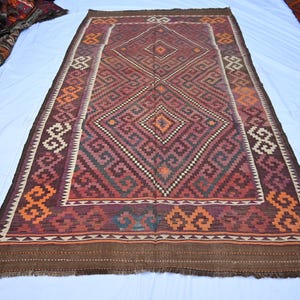 Puede incluir: Una alfombra tejida rectangular con un motivo central de diamantes en tonos rojo oscuro, marrón y naranja. La superficie muestra patrones geométricos intrincados y un diseño de borde repetitivo con flecos en los extremos superior e inferior.