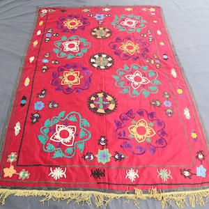May include: A vibrant red textile featuring large, embroidered floral designs in white, yellow, green, purple, and blue. The edges are trimmed with a gold fringe. This decorative textile is likely a rug or wall hanging.