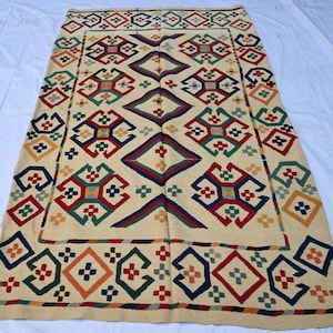 May include: Rectangular beige rug with a geometric pattern in red, green, blue, and orange. The rug features diamond and square shapes, creating a symmetrical design. The border has a repeating pattern of squares and diamonds.