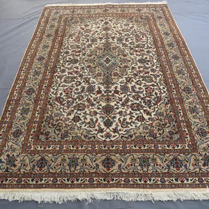 May include: Rectangular beige rug with an intricate floral pattern in shades of red, blue, and brown. The rug has a detailed border and a central medallion design. The rug has a fringe on the short ends.