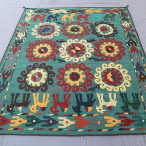 May include: A rectangular teal rug featuring a central pattern of nine large circular floral motifs in red, yellow, and cream. The border displays rows of stylized camels and birds in various colors including red, yellow, blue, and dark brown.