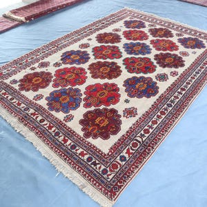 May include: A white rug with a traditional Persian design. The rug features a repeating pattern of red, blue, and yellow flowers on a white background. The rug has a fringed border.