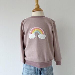 Könnte beinhalten: Ein mauvefarbener Pullover mit einem Regenbogen- und Wolken-Applikation. Der Regenbogen ist aus Pastellfarben und die Wolken sind weiß.