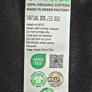 May include: A white care label with green text and symbols. The label reads "Verdant Universe 100% Organic Cotton Made in Green Factory". It also includes washing instructions, a QR code, and several certifications including OEKO-TEX, GOTS, WRAP, and Carbon Neutral.
