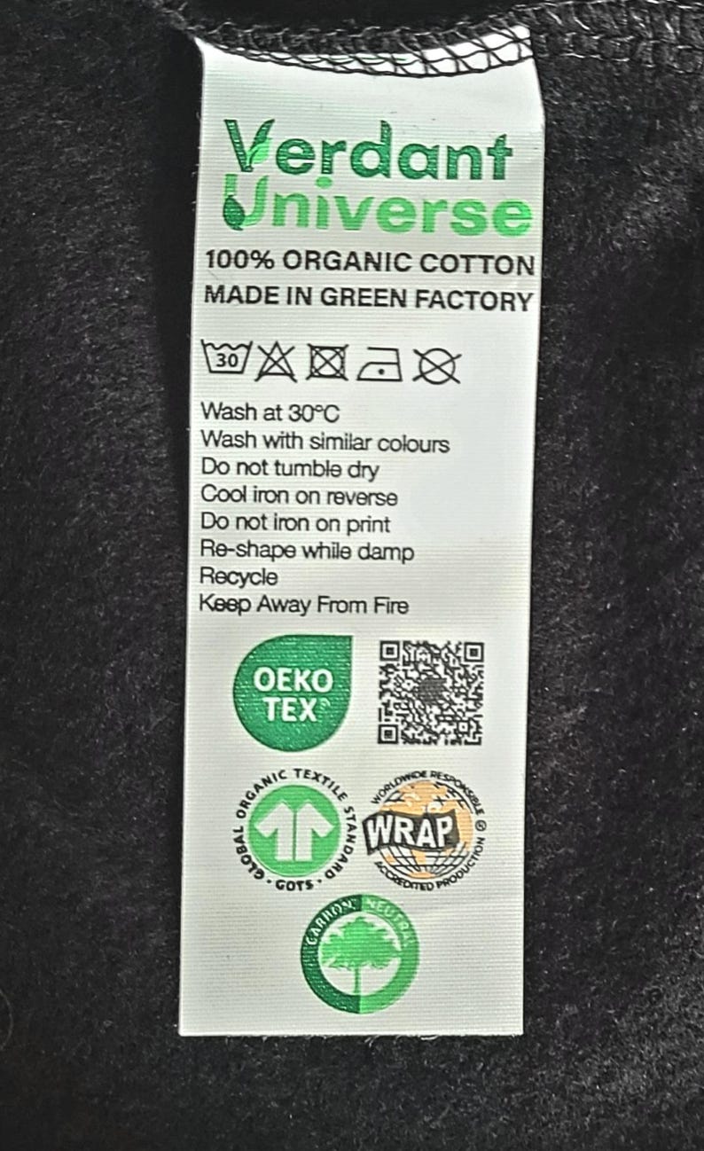 May include: A white care label with green text and symbols. The label reads "Verdant Universe 100% Organic Cotton Made in Green Factory". It also includes washing instructions, a QR code, and several certifications including OEKO-TEX, GOTS, WRAP, and Carbon Neutral.