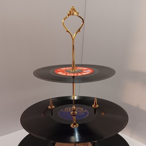May include: A three-tiered cake stand made from black vinyl records with a gold metal handle. The stand is decorated with gold accents.