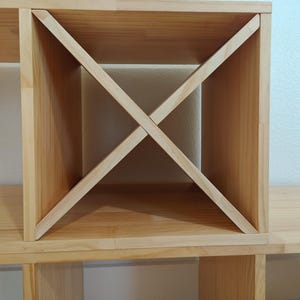 May include: A light-colored wooden storage unit featuring a square compartment divided by two intersecting diagonal boards that form an X-shaped pattern. The structure is constructed from smooth, natural-toned wood planks with visible grain.