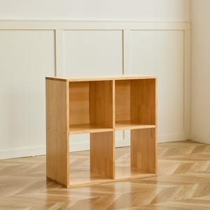 May include: A light-colored wooden cube shelf unit with four open compartments. The unit is made of light-colored wood and sits on a wooden floor. The background is a white wall.