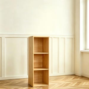 May include: A three-shelf wooden bookcase with a natural finish. The bookcase is rectangular and stands upright, with three evenly spaced shelves. The wood grain is visible. The background is a light-colored wall and a wooden floor.