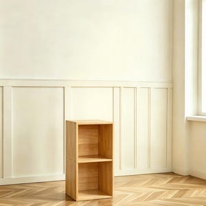 May include: A two-shelf wooden bookcase in a light wood finish. The bookcase is rectangular with two open compartments. It stands on a herringbone patterned wood floor, against a cream-colored wall.