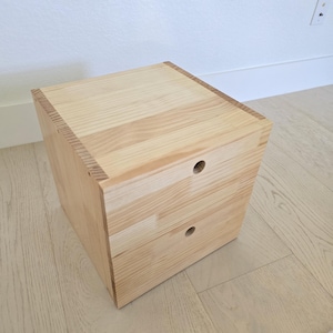 May include: A light-colored wooden cube-shaped storage unit with two drawers. The unit is constructed from light-colored wood planks and features two circular drawer pulls. The wood grain is visible, and the unit sits on a light-colored floor.