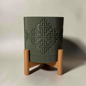 May include: A cylindrical olive green planter featuring a textured, geometric relief pattern on its exterior surface. The planter is supported by a light brown, three-legged stand with a cross-shaped base, providing elevation for the container.