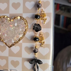 May include: A gold-toned phone charm with a black rose, beads, and a bow. The charm is attached to a phone case with a heart-shaped design. The charm has a heart-shaped gold clasp.