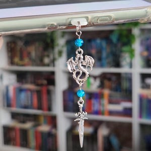 May include: A silver charm featuring a dragon and sword, with blue bead accents, attached to a clear phone case. The design suggests a fantasy or book-related theme.