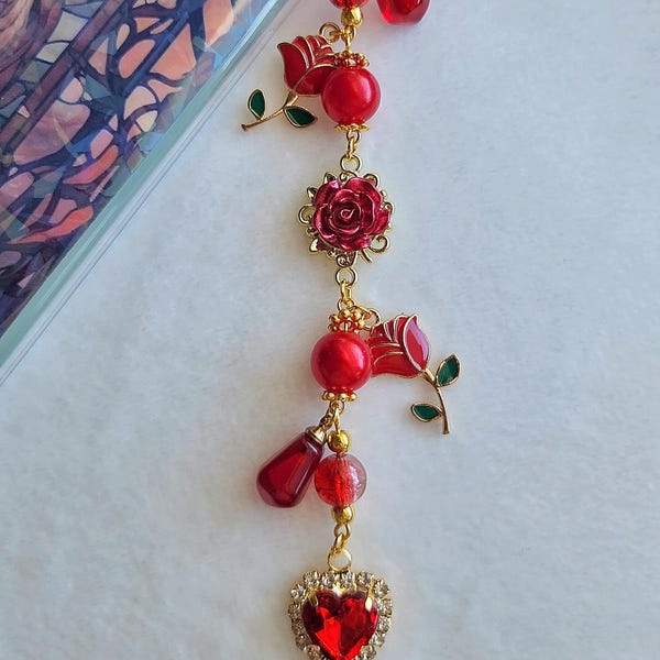 Puede incluir: Un charm de teléfono dorado con detalles de rosas rojas y en forma de corazón. El charm presenta cuentas rojas, colgantes en forma de rosa y un colgante en forma de corazón con una gema roja rodeada de cristales transparentes.
