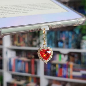 May include: A red heart-shaped charm with a crystal border, suspended from a gold chain. The charm is attached to a tablet. The background features a bookshelf filled with books of various colours.