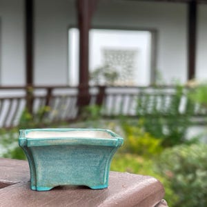 May include: A rectangular ceramic bonsai pot with a turquoise blue glaze and a white interior. The pot features a flared rim, slightly tapered sides, and four small feet at the base, showcasing a smooth, glossy finish with subtle color variations.