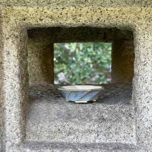 May include: A small, shallow ceramic bowl with a hexagonal rim and a faceted exterior. The glaze features a blend of soft blue and muted grey tones, transitioning into a creamy off-white interior. The bowl rests on a small, raised base.