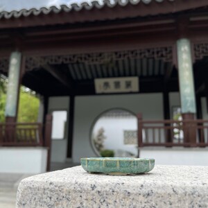 May include: A small, shallow, scalloped ceramic dish with a mottled teal and green glaze. The dish features a series of vertical ridges along its exterior edge and rests on a light-colored, textured stone surface.
