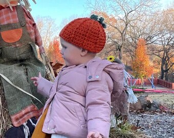 May include: A child wearing an orange crochet pumpkin hat with a green stem and a light pink jacket. The child is standing near a scarecrow and hay bale, with trees in the background.