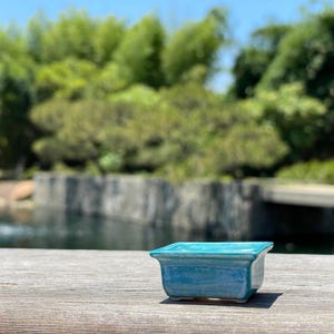 Puede incluir: Pequeño tiesto cuadrado de cerámica turquesa con acabado brillante. La maceta tiene un borde ligeramente ensanchado y se apoya sobre una superficie de madera, con un fondo borroso de vegetación y agua.