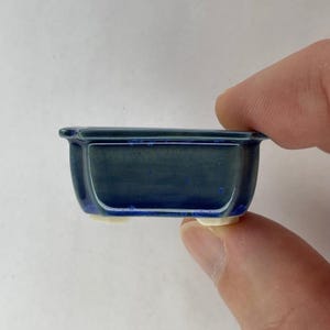 May include: A small, rectangular bonsai pot with a dark blue glaze. The ceramic pot has rounded corners and sits on four small, white feet. The pot is held by a person's fingers.
