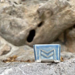 May include: A small, square ceramic pot with a light blue and white glazed finish. The exterior features a geometric design with two horizontal V-shaped indentations on the front face. The pot has a flat base and four short, sturdy feet.