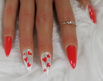 May include: Close-up of a hand with red and silver nail polish. The silver nails have red heart designs. The ring finger has a thin gold band with small clear stones.