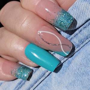 Peut inclure: Une main montrant de longs faux ongles carrés. Les ongles présentent une base rose clair avec un dégradé de paillettes turquoise, un ongle turquoise uni et un ongle avec un motif tourbillon blanc et argenté. Les ongles sont posés sur un fond en jean.