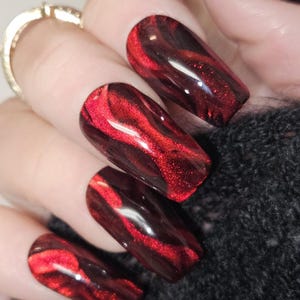 Può includere: Primo piano di nail art rossa e nera. Le unghie sono di un rosso intenso con motivi neri a spirale e una finitura lucida. Il design è impreziosito da glitter, creando un effetto scintillante. Le unghie sono lunghe e rettangolari.