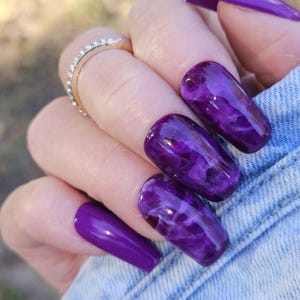 May include: Close-up of a hand with long, purple press-on nails. The nails have a marbled design with shades of purple and white. A gold ring with small, clear stones is on the ring finger. The hand is resting on a denim fabric.