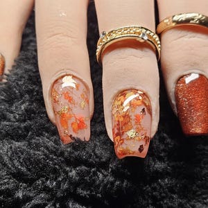 May include: Manicured nails featuring a mix of solid copper glitter polish and clear base coats embedded with orange leaf-like flakes and gold foil accents. Two gold-toned metal rings are worn on the fingers, one with a textured band and one plain.