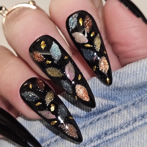 May include: Long, pointed, black fingernails with a glossy finish. Each nail has a design of colourful, glittery leaf shapes and gold flecks. The nails are set against a denim background.