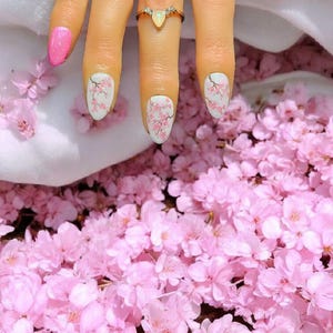 May include: Close-up of a hand with oval-shaped nails featuring a cherry blossom design. The nails are painted white with pink cherry blossoms. The hand is wearing a gold ring with a clear stone.