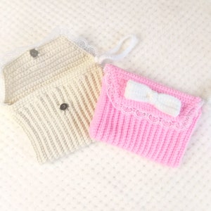 May include: Two handmade crochet pouches. One is ivory with a magnetic snap closure and lace trim. The other is pink with a white bow and lace trim. Both have a ribbed texture and a white handle.