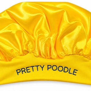 May include: A bright yellow satin bonnet featuring a gathered, rounded top and a wide, flat headband. The front of the headband is embroidered with the text PRETTY POODLE in dark blue capital letters, creating a distinct contrast against the yellow.