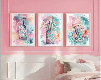 Op de afbeelding: Set van drie ingelijste aquarelprints. Elke print toont een ander kunstbenodigdheid: een verftube, penselen in een decoratieve houder en een verfpalet. De prints hebben een kleurrijke, abstracte achtergrond met een roze en blauwgroene kleurstelling.