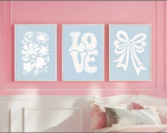 Op de afbeelding: Drie ingelijste kunstdrukken aan een roze muur. De prints tonen witte ontwerpen op een lichtblauwe achtergrond. Eén print heeft een bloemenontwerp, een andere spelt "LOVE" en de derde toont een strik.
