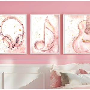 Könnte beinhalten: Drei gerahmte Aquarelldrucke mit musikalischen Themen. Die Drucke zeigen Kopfhörer, eine Musiknote und eine Gitarre, alle in sanften Rosatönen mit goldenen Akzenten. Das Kunstwerk ist vor einer rosa Wand platziert.