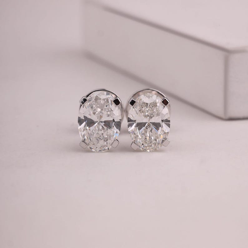 May include: A pair of oval-cut diamond stud earrings. The diamonds are set in a silver metal. The earrings are displayed on a white surface. The diamonds sparkle and reflect light.