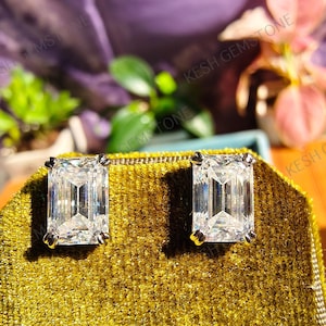 May include: A pair of emerald-cut diamond stud earrings. The clear, rectangular gemstones are set in gold-colored prongs. The earrings are displayed on a gold velvet surface, with the text "KESH GEMSTONE" visible in the background.