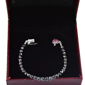 Könnte beinhalten: Ein silbernes Armband mit dunkelgrauen facettierten Perlen und einem großen, ovalen, rosa Edelstein. Das Armband wird in einer roten Schmuckschatulle mit schwarzem Innenraum präsentiert.